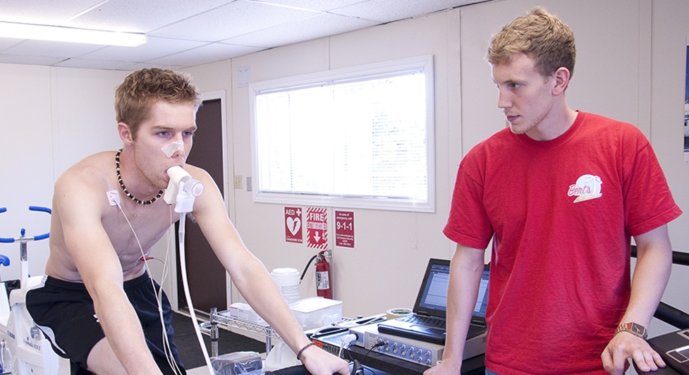 Young shirtless man wearing sensors pedaling a cycle ergometer while a lab technician in a red shirt monitors data