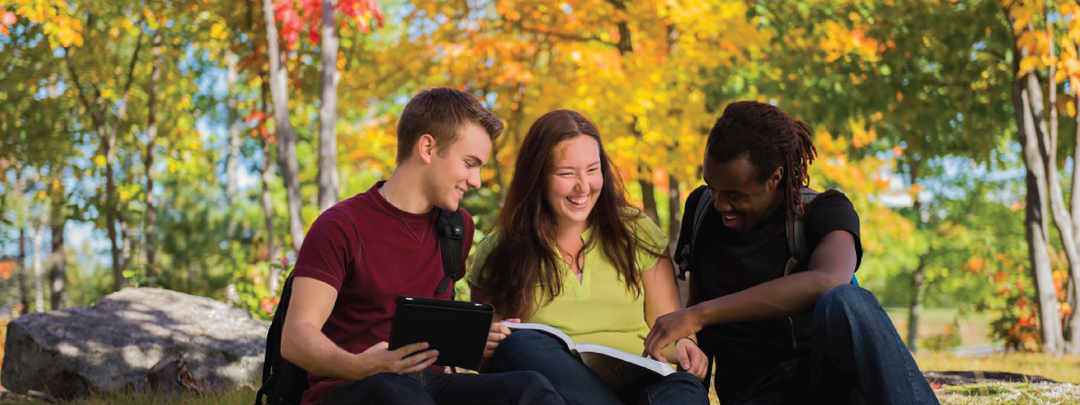 3 students sitting on the grass chatting with the fall background behind them