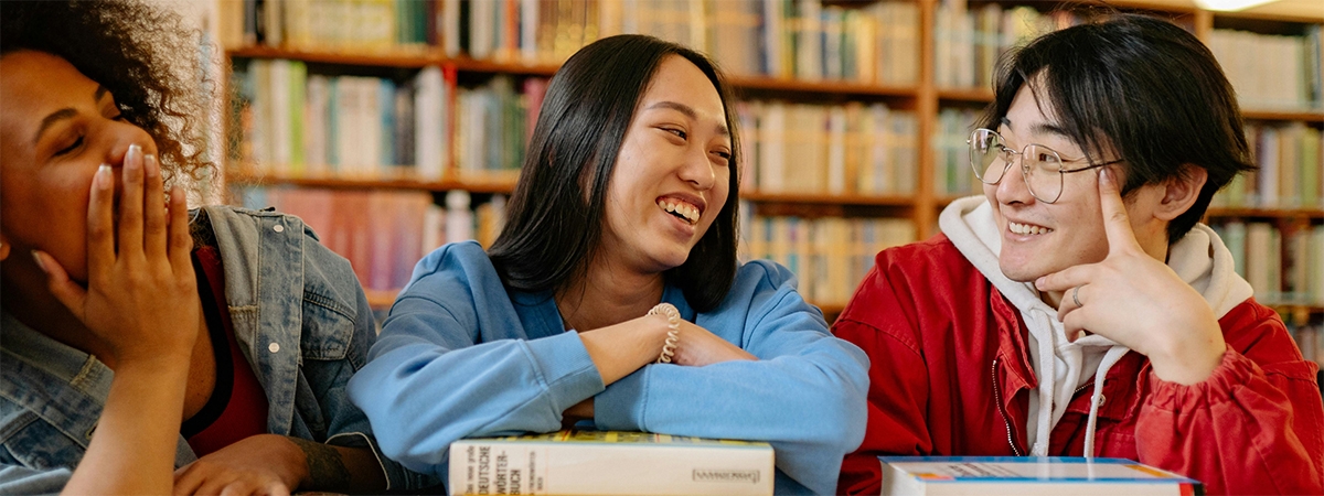 Three students sitting together laughing with a wall of library books behind them.