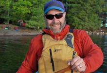 Terry Dubroy smiles at the camera from his canoe with a forested background.